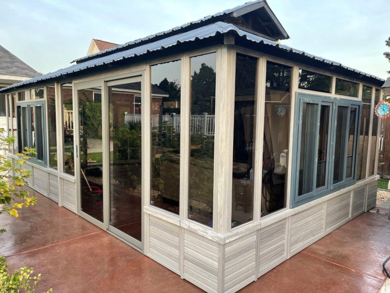 A gray, glass-enclosed sunroom with a corrugated roof on a concrete patio.