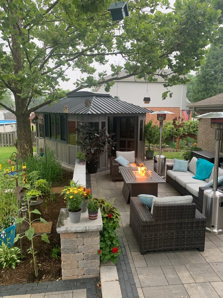 Backyard patio with wicker furniture, fire pit, planters, lush garden, and a gazebo under a large tree.