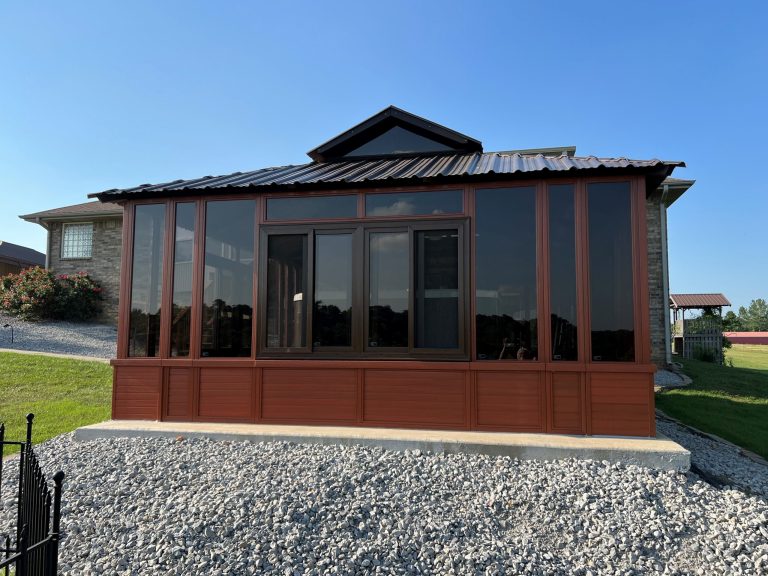 A brown glass sunroom with a metal roof, surrounded by gravel and a grassy area, attached to a brick house.