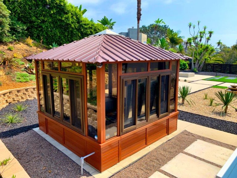 A wooden gazebo with glass windows, situated in a landscaped garden with plants and a tiled walkway.