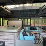 Enclosed patio with a hot tub, turquoise sectional sofa, black and white rug, surrounded by large windows and forest view.
