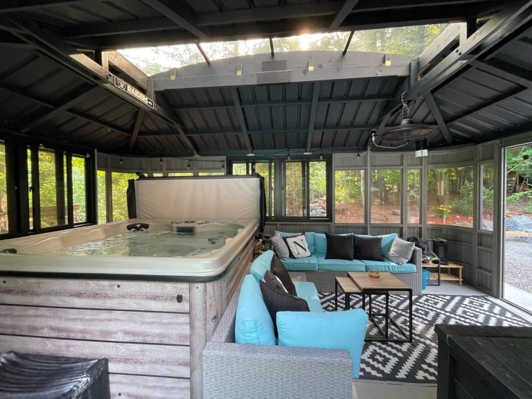 Enclosed patio with a hot tub, turquoise sectional sofa, black and white rug, surrounded by large windows and forest view.