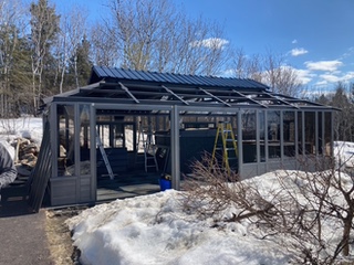 A partially constructed greenhouse with a blue roof, surrounded by snow, and a ladder inside under a clear blue sky.