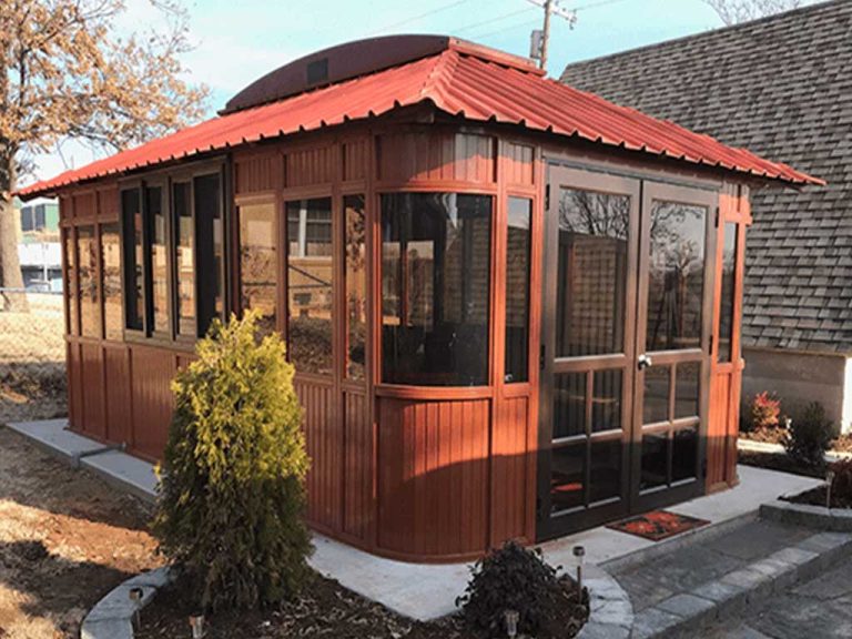 Red-roofed wooden gazebo with glass walls, set on concrete, surrounded by small plants and trees.