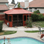 A backyard with a swimming pool, a small round table and two chairs, green bushes, neighboring houses in the background, and an Aspen 12x14 gazebo on sale.