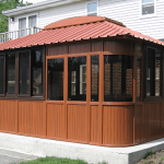 The Aspen 12x16 is a brown wooden outdoor gazebo with large windows and a red metal roof, set on a concrete patio beside a brick and white-sided house—ideal as a hot tub gazebo or swim spa enclosure, surrounded by grass and pavement.