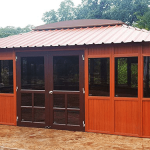 Redwood gazebo with glass doors and windows, set on a concrete patio, surrounded by lush greenery.