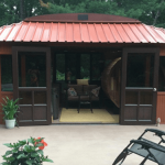 Red-roofed shed with open doors, surrounded by chairs, plants, a fire pit, and trees in the background.