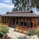 Wooden gazebo with glass windows and a metal roof, surrounded by a garden with shrubs, overlooking a distant view.