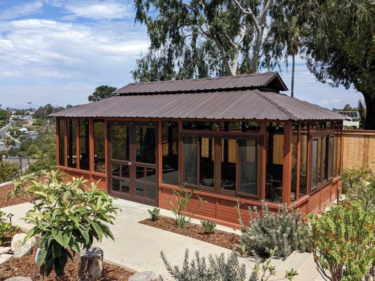 Wooden gazebo with glass windows and a metal roof, surrounded by a garden with shrubs, overlooking a distant view.