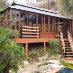 Wooden cabin with large windows on a hillside, surrounded by plants, with stairs leading to a stone path near a pool.
