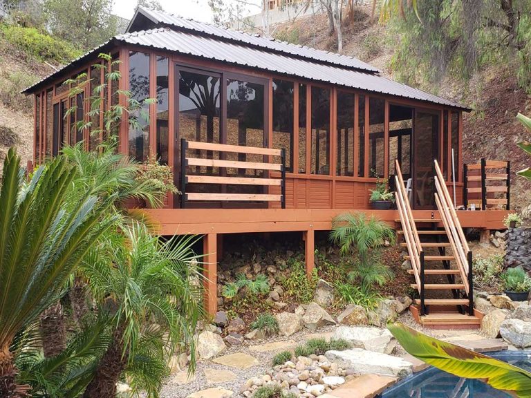 Wooden cabin with large windows on a hillside, surrounded by plants, with stairs leading to a stone path near a pool.