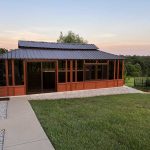 A wooden gazebo with large windows and a metal roof on a concrete and gravel patio, surrounded by grassy landscape.