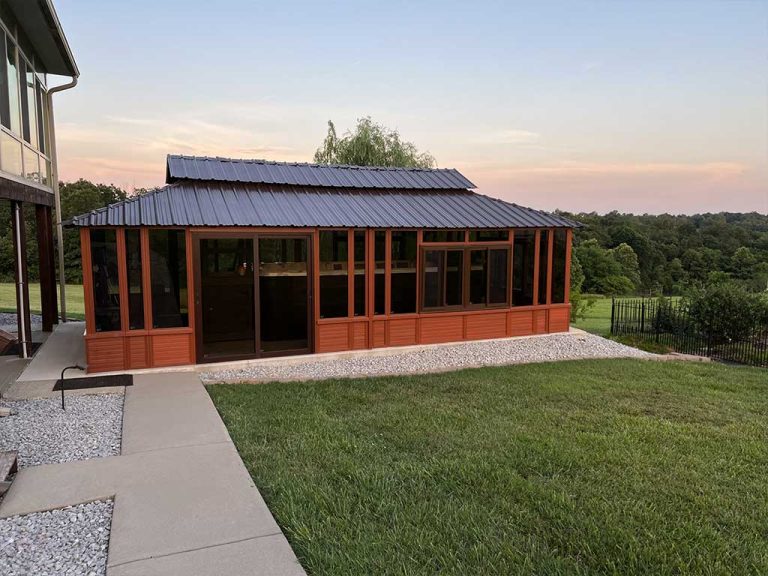 A wooden gazebo with large windows and a metal roof on a concrete and gravel patio, surrounded by grassy landscape.
