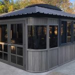 Gray gazebo with large windows and a metal roof on a concrete patio surrounded by trees.