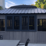 Gray outdoor gazebo with black-framed windows on a patio next to garden greenery.