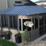 A small wooden gazebo with a metal roof, surrounded by plants and hanging flower baskets, set in a backyard.