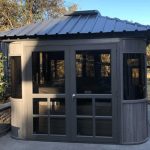 Small gray wooden gazebo with glass windows and metal roof, set on a concrete patio with trees and fence in the background.