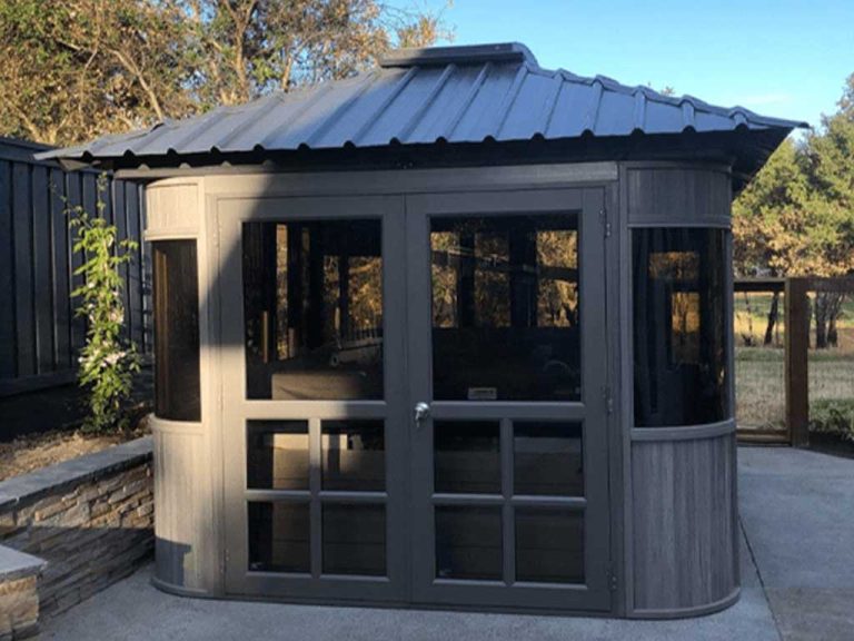Small gray wooden gazebo with glass windows and metal roof, set on a concrete patio with trees and fence in the background.