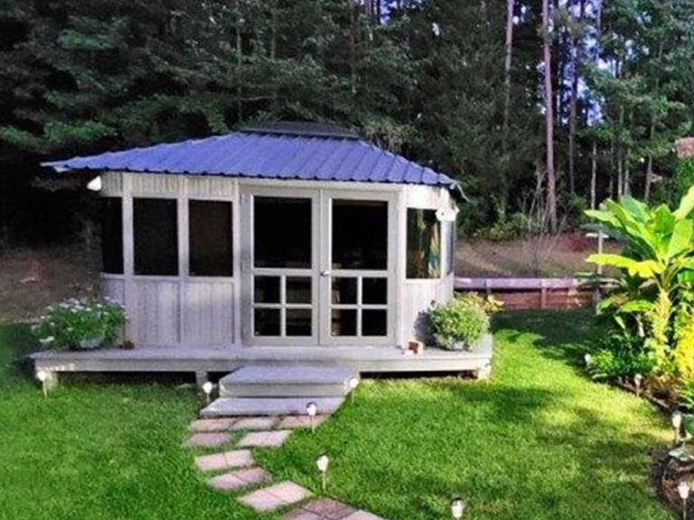 A small wooden gazebo with a blue metal roof on a grassy lawn surrounded by trees.