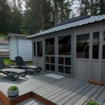 Deck with two loungers, small porch, and potted plants, next to a gray building surrounded by trees.