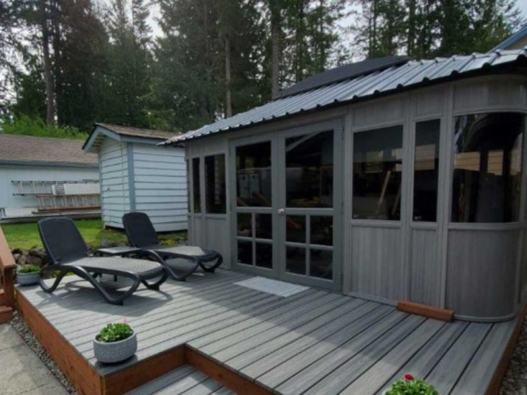 Deck with two loungers, small porch, and potted plants, next to a gray building surrounded by trees.