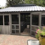 Outdoor spa surrounded by a grey gazebo with open doors, brick patio, and potted plants nearby.