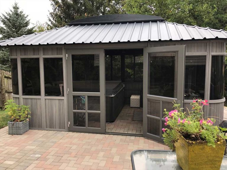 Outdoor spa surrounded by a grey gazebo with open doors, brick patio, and potted plants nearby.