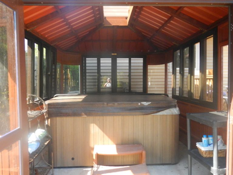 Enclosed gazebo with a hot tub, steps, shelves, and a view of wooden exterior siding through large windows.