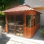 Wooden gazebo with glass doors and windows attached to a house, surrounded by plants and patio bricks.