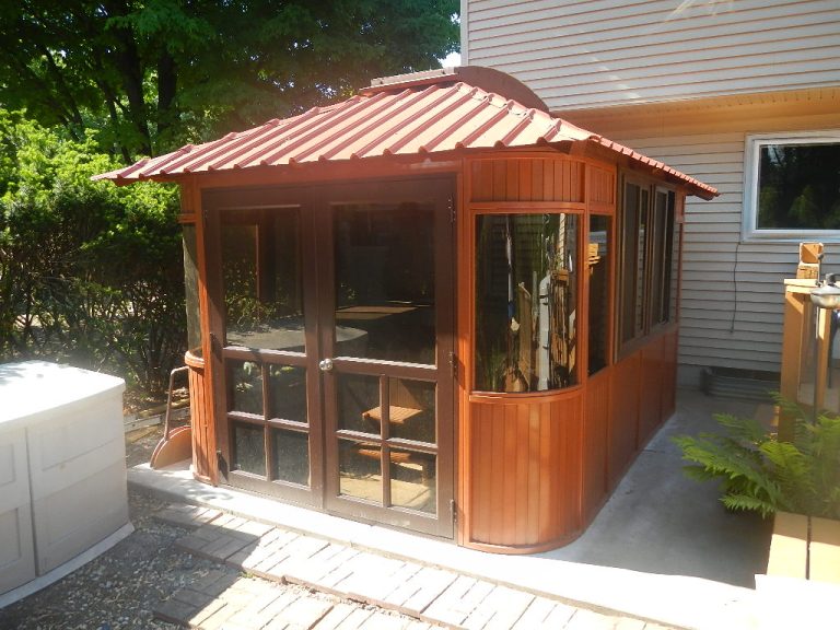 Wooden gazebo with glass doors and windows attached to a house, surrounded by plants and patio bricks.