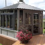 Small glass-enclosed sunroom with metal roof on wooden deck, surrounded by grass and a potted pink flower.