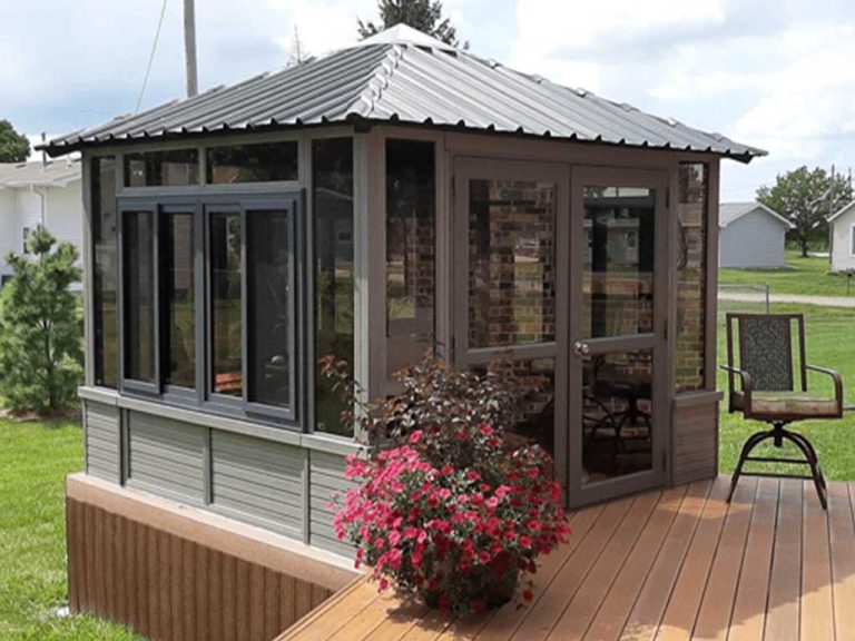 Small glass-enclosed sunroom with metal roof on wooden deck, surrounded by grass and a potted pink flower.