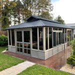 Glass-enclosed garden gazebo with a metal roof, surrounded by lush greenery and trees.