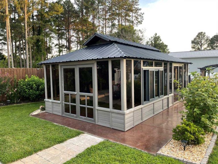 Glass-enclosed garden gazebo with a metal roof, surrounded by lush greenery and trees.