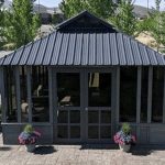Gray garden gazebo with glass panes, surrounded by potted flowers and trees on a stone patio.