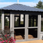 A gray gazebo with a metal roof on a wooden deck, surrounded by green grass and vibrant pink flowers.
