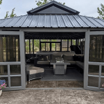 Gray gazebo with open doors, cozy couches, a table inside, and potted flowers outside.
