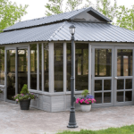 A glass-enclosed patio with a metal roof, surrounded by potted flowers and trees, on a stone-paved surface.