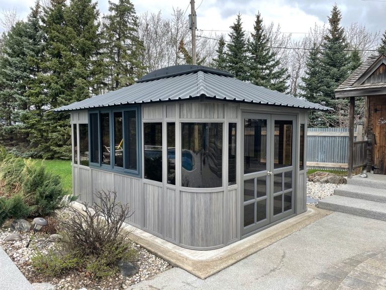 A gray gazebo with large windows sits on a paved area surrounded by evergreen trees and a small garden.