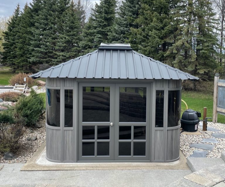 Gray outdoor gazebo with dark windows, metallic roof, surrounded by trees and garden stones.