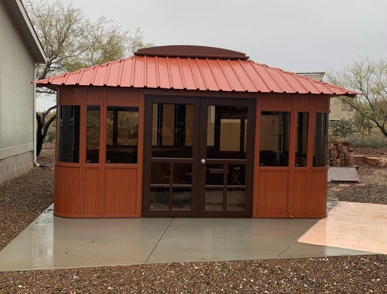 Small wooden gazebo with a red metal roof on concrete, near a building and trees.