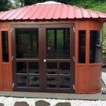 The Aspen 12x12 is a brown wooden gazebo with a red metal roof, set on a stone patio surrounded by white gravel, garden decor, and shrubs—an inviting upgrade for any outdoor gazebos or pergolas collection.
