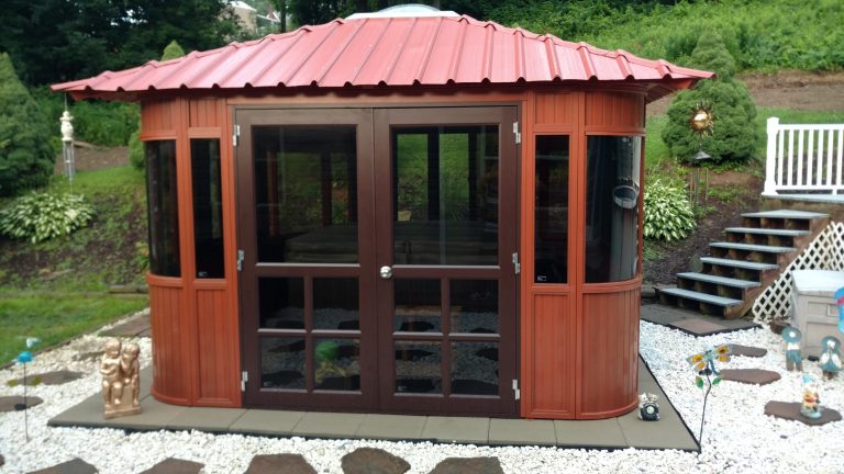The Aspen 12x12 is a brown wooden gazebo with a red metal roof, set on a stone patio surrounded by white gravel, garden decor, and shrubs—an inviting upgrade for any outdoor gazebos or pergolas collection.