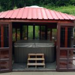 Gazebo with red roof and open doors revealing a covered hot tub inside, surrounded by a garden and decorative items.