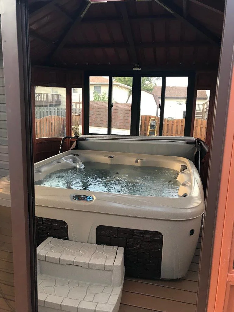 Indoor hot tub with bubbling water in a wooden gazebo, steps leading up, and windows overlooking a fenced backyard.