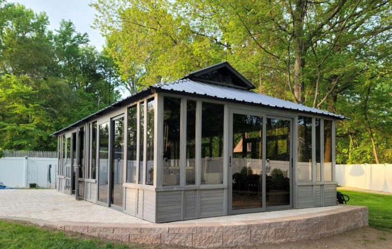 Glass-enclosed pavilion with metal roof in a backyard, surrounded by trees and a white fence.