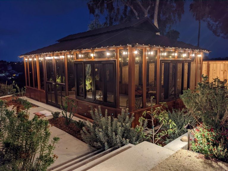 Glass-walled house at night with string lights, surrounded by plants and steps leading to it.