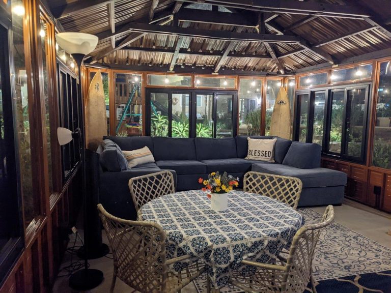 Cozy sunroom with a blue sectional sofa, round table with floral cloth, wicker chairs, surfboards, and bright lighting.