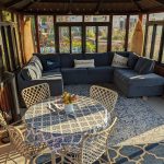 Sunlit sunroom with a blue sectional sofa, patterned rug, round table with floral tablecloth, and wicker chairs.